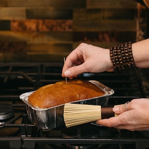 May include: A freshly baked loaf of golden-brown bread in a disposable aluminum baking pan. A hand is inserting a toothpick to check for doneness. A bamboo whisk is held in the other hand, ready for use. The scene is set on a gas stovetop.