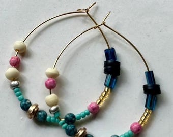 May include: A pair of gold-toned hoop earrings decorated with an assortment of beads. The beads include cream-colored spheres, pink marbled spheres, turquoise marbled spheres, translucent blue cylinders, black discs, and small metallic spacers.