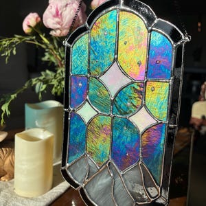 May include: Stained glass window panel with a black metal frame. The glass features iridescent colors of blue, green, and yellow, with white and pink accents. The panel is hanging from a chain. Decorative home decor.