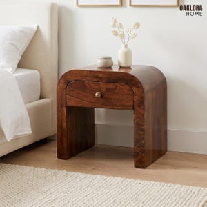 May include: A dark brown wooden nightstand with a curved waterfall edge design and a single drawer featuring a small round metal knob. The nightstand is positioned next to a bed and holds a small white ceramic vase with dried flowers.