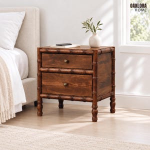 May include: A dark brown wooden nightstand with two drawers, featuring a bamboo-style frame and small round brass-colored knobs. The nightstand has four legs and a smooth finish, with a white ceramic vase containing green leaves on top.