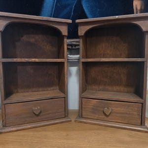 May include: A pair of matching dark brown wooden wall shelves, each featuring an arched top, two open storage compartments, and a single bottom drawer with a small heart-shaped wooden knob centered on the front panel of each drawer.