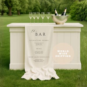 Könnte beinhalten: Eine weiße Outdoor-Bar mit einem cremefarbenen Leinenbanner mit der Aufschrift "The Bar". Die Bar hat eine silberne Schüssel mit zwei Flaschen und sechs Sektgläsern. Die Bar steht auf einer grünen Wiese mit Bäumen im Hintergrund.