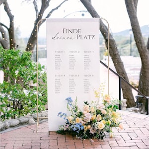 Puede incluir: Un plano de asientos de boda blanco con marco dorado. El gráfico muestra los nombres de los invitados organizados por número de mesa. El texto "FINDE deinen PLATZ" está en la parte superior. Un arreglo floral de flores amarillas, azules y rosas está en la base.