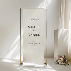 May include: A wedding welcome sign with gold-colored metal frame. The white banner displays the names "Sophie & Daniel" with the wedding date and location. A small floral arrangement sits at the base of the sign.