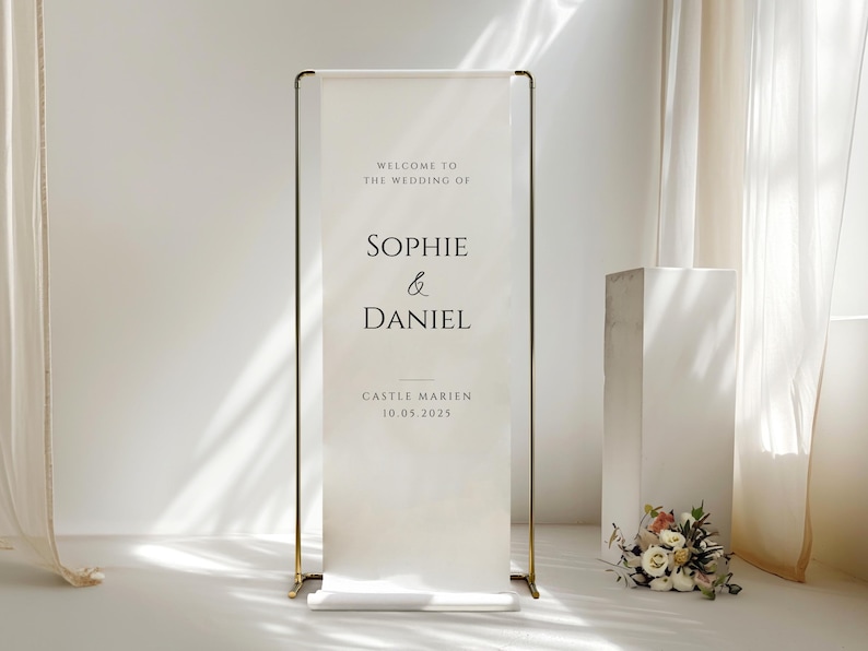 May include: A wedding welcome sign with gold-colored metal frame. The white banner displays the names "Sophie & Daniel" with the wedding date and location. A small floral arrangement sits at the base of the sign.