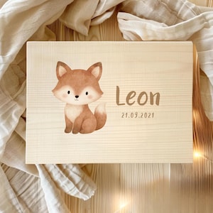 Pode incluir: Uma caixa de madeira de cor clara com uma ilustração em aquarela de uma raposa. A raposa é marrom e branca. O nome "Leon" e a data "21.09.2021" são impressos em marrom na caixa.