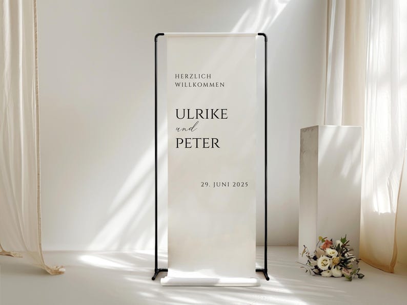 Puede incluir: Un cartel de bienvenida blanco con texto negro en un soporte negro. El cartel dice "Herzlich Willkommen Ulrike und Peter 29. Juni 2025".