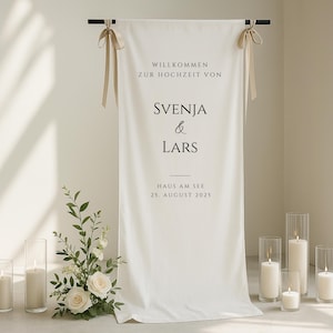 May include: A white wedding banner with black text that reads "Willkommen zur Hochzeit von Svenja & Lars Haus am See 25. August 2025." The banner is decorated with beige ribbons and is displayed with candles and flowers.