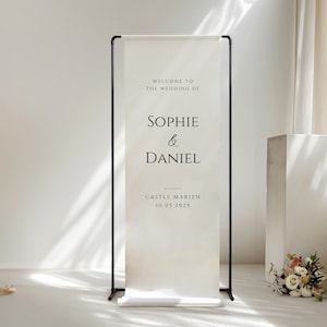 May include: A wedding welcome sign with black frame and a cream-colored banner. The banner reads "Welcome to the wedding of Sophie & Daniel" and includes the date and location.