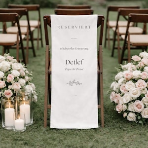Pode incluir: Um banner de tecido branco com a palavra alemã "RESERVIERT" numa cadeira de madeira. O banner inclui texto, detalhes florais e está rodeado por cadeiras, flores e velas, sugerindo um casamento ou um memorial.