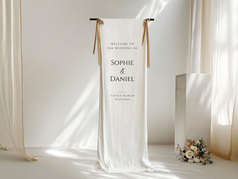 May include: A white wedding welcome sign with the text "WELCOME TO THE WEDDING OF SOPHIE & DANIEL" and the date "10.05.2023". The sign is suspended from a dark rod with gold ribbons. A small floral arrangement is at the base.