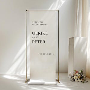 Puede incluir: Un cartel de bienvenida blanco con marco dorado, con el texto "Herzlich Willkommen" (alem&aacute;n para "Bienvenido"), "Ulrike und Peter" (alem&aacute;n para "Ulrike y Peter") y "29. Juni 2025" (alem&aacute;n para "29 de junio de 2025").