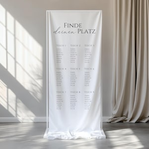Könnte beinhalten: Ein weißes Hochzeits-Sitzplan aus Stoff mit schwarzem Text. Der Text lautet "FINDE deinen PLATZ" oben, gefolgt von Tischnummern und Gästenamen. Der Plan wird auf einem weißen Metallrahmen angezeigt.