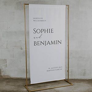 Puede incluir: Un soporte de metal dorado con una pancarta blanca que dice "Herzlich Willkommen Sophie und Benjamin 14. August 2023 Marthas Garten".