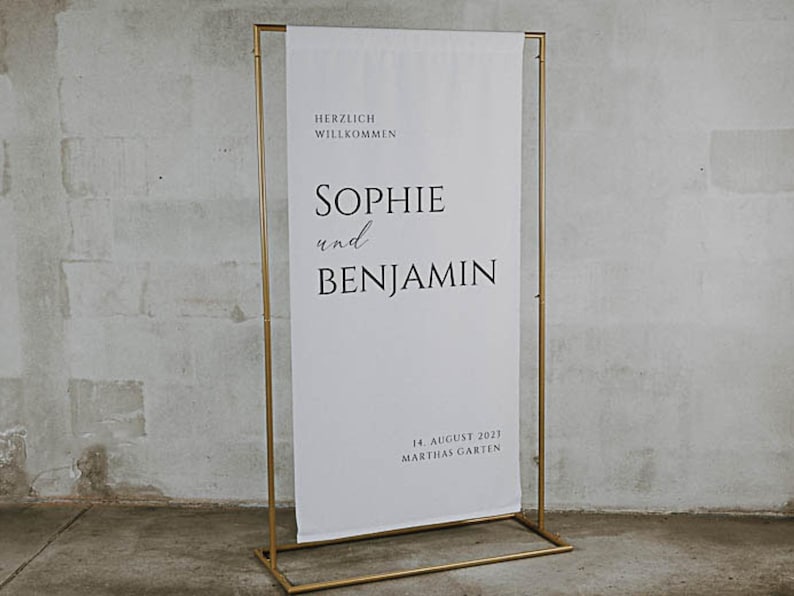 Puede incluir: Un soporte de metal dorado con una pancarta blanca que dice "Herzlich Willkommen Sophie und Benjamin 14. August 2023 Marthas Garten".