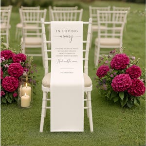 Könnte beinhalten: Weißer Stuhl mit cremefarbenem Sitz, über den ein weißes Banner mit der Aufschrift "IN LOVING memory" und deutschem Text drapiert ist. Pinke Blumen und Kerzen flankieren den Stuhl auf einer grünen Wiese.