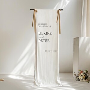 Puede incluir: Tel&oacute;n de fondo de tela blanca con lazos de cinta marr&oacute;n, un letrero que dice "Herzlich Willkommen Ulrike und Peter 29. Juni 2025".