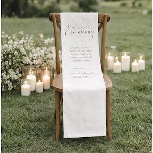 Könnte beinhalten: Ein weißes Stuhlsash mit schwarzer Schrift, die „In liebevoller Erinnerung“ und eine Nachricht darunter lautet. Das Sash ist über einen Holzstuhl drapiert, mit Kerzen und Blumen im Hintergrund.
