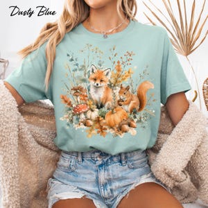 May include: A dusty blue short-sleeve t-shirt featuring a graphic print of a fox and a squirrel surrounded by autumn elements including pumpkins, mushrooms, acorns, and foliage. The text Dusty Blue is printed in the upper left corner of the image.