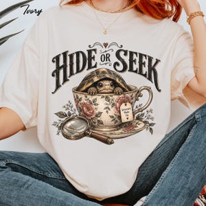 May include: An ivory t-shirt featuring a graphic of a turtle inside a floral teacup. The text reads HIDE OR SEEK and Introvert Mode On. A magnifying glass is positioned below the teacup. The design includes roses and leaves in muted tones.