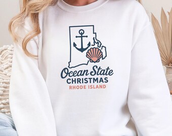 May include: White sweatshirt featuring a graphic of the Rhode Island outline with an anchor and seashell. The text reads "Ocean State Christmas Rhode Island" in navy and coral. The word "WHITE" is at the bottom.