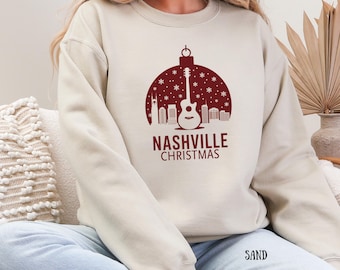May include: Sand-coloured sweatshirt with a maroon graphic of a guitar inside a Christmas ornament, with the text "Nashville Christmas" below. The design includes snowflakes and city skyline silhouettes. The word "SAND" is printed at the bottom.