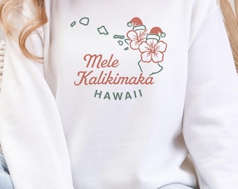 May include: White sweatshirt with a Hawaiian Christmas design. The design includes the text "Mele Kalikimaka HAWAII" in red and green, along with illustrations of flowers and Santa hats. The word "WHITE" is printed at the bottom.