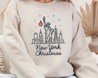 May include: Sand-colored sweatshirt with a graphic of the New York City skyline, the Statue of Liberty, snowflakes, and a Christmas ornament. The text "New York Christmas" is printed below the graphic.