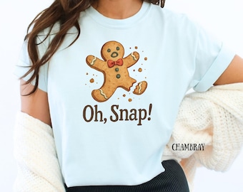 May include: Pale blue t-shirt with a gingerbread man graphic, a red bow tie, and the phrase "Oh, Snap!". The gingerbread man has a broken leg. The word "CHAMBRAY" is printed on the right side of the shirt.