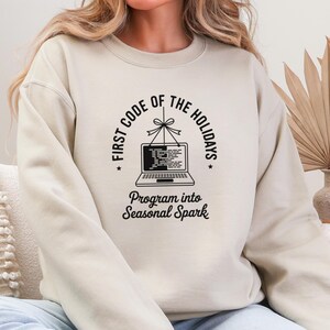 May include: A cream-coloured sweatshirt featuring a black graphic. The design includes the text "FIRST CODE OF THE HOLIDAYS" encircling a laptop illustration. Beneath the laptop, it reads "Program into Seasonal Spark".