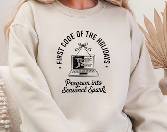 Puede incluir: Sudadera beige con un diseño negro. El diseño circular dice "FIRST CODE OF THE HOLIDAYS" alrededor de la imagen de un portátil. Debajo del portátil, el texto dice "Program into Seasonal Spark".