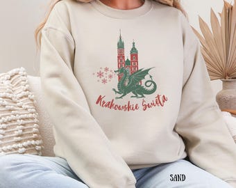 Może przedstawiać: Beżowa bluza z zielonym smokiem i czerwoną i zieloną ilustracją budynku. Tekst "Krakowskie Swieta" jest czerwony. Słowo "SAND" jest wydrukowane na dole w kolorze czarnym.