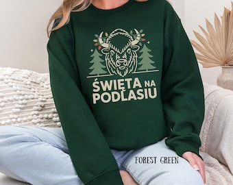 Może przedstawiać: Zielona bluza w kolorze leśnej zieleni z grafiką przedstawiającą bizona i białym napisem "ŚWIĘTA NA PODLASIU". Bizon jest ozdobiony elementami świątecznymi. Bluza jest w jednolitym kolorze i ma okrągły dekolt.