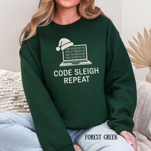 Puede incluir: Sudadera verde bosque con el texto blanco "CODE SLEIGH REPEAT". El diseño incluye un portátil con código binario y un gorro de Papá Noel. Las palabras "FOREST GREEN" están impresas en la parte inferior.