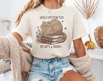May include: A cream-coloured t-shirt with the text "EASILY DISTRACTED BY CATS & BOOKS." The graphic shows a brown cat on a stack of books, with flowers and a ladybug. The shirt is a casual, comfortable style.