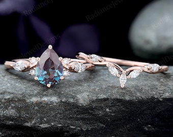 May include: A rose gold engagement ring set featuring a pear-shaped, color-changing gemstone and a matching band. The rings are adorned with small, clear accent stones and are displayed on a dark, textured surface. The text "Radiance Jewel Designs" is visible.