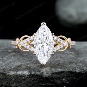 Puede incluir: Un anillo de compromiso con diamantes de talla marquesa y una banda de oro. El anillo presenta un gran diamante transparente de talla marquesa como pieza central, rodeado de diamantes más pequeños y detalles intrincados en oro. El anillo está sobre una superficie oscura y texturizada.