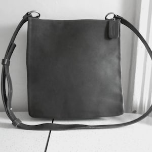 May include: A dark gray leather crossbody bag with a long adjustable strap. The bag has a rectangular shape with a small tag attached to the side. The strap is attached to the bag with silver-colored metal rings.