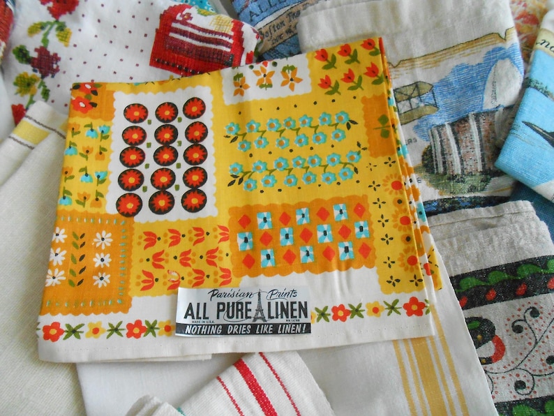 May include: A collection of vintage linen textiles with colorful floral and geometric patterns. The focal point is a yellow linen with orange, red, and blue designs. The text "ALL PURE LINEN" is visible. Other linens feature various patterns.