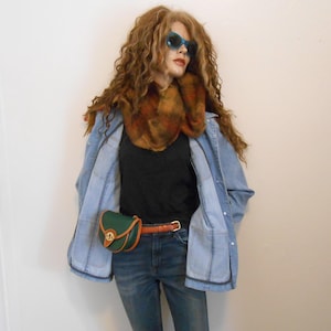 May include: A mannequin wearing a denim jacket, black shirt, and jeans. Accessories include a green and brown fanny pack, a brown scarf, a brown belt, and blue sunglasses. The mannequin has long, curly auburn hair.
