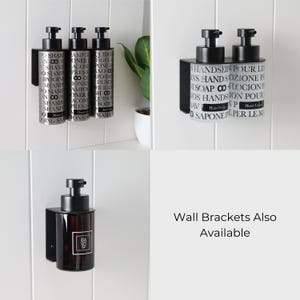 May include: Black metal wall brackets holding various soap and shampoo dispensers. The dispensers are cylindrical with black pump tops and labels with text. The image also shows a plant in a white pot.