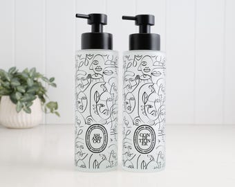 May include: Two cylindrical frosted glass bottles with black pump dispensers. Each bottle features a black line art pattern of abstract faces and a circular label. One bottle is labeled SHAMPOO and the other is labeled CONDITIONER.
