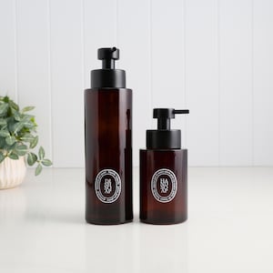 May include: Two brown glass soap dispensers with black pumps. The taller dispenser is labelled "Dish Soap", and the smaller one is labelled "Hand Soap". Both have white circular labels with text. The dispensers are on a white surface.