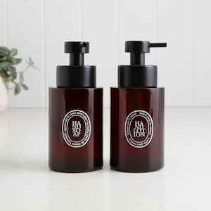 Könnte beinhalten: Zwei braune Glasflaschen mit schwarzen Pumpen. Auf einer steht "Hand Soap" und auf der anderen "Hand Lotion" in einem weißen, kreisförmigen Design. Die Flaschen sind zylindrisch und stehen auf einer weißen Oberfläche.