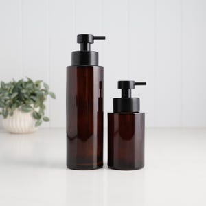 May include: Two brown glass soap dispensers with black pump tops. The larger dispenser is taller than the smaller one. Both dispensers are cylindrical and sit on a white surface. A small plant in a white pot is in the background.