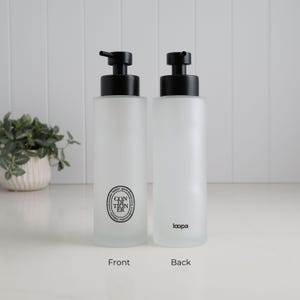 Front and reverse view of frosted glass conditioner dispenser with matte black pump, featuring minimalist circular label on one side and subtle branding on the other. Refillable bathroom bottle with soft matte finish, designed for clean, modern shower organisation and a calm, uncluttered aesthetic.