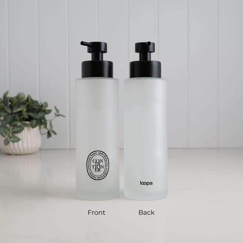 Front and reverse view of frosted glass conditioner dispenser with matte black pump, featuring minimalist circular label on one side and subtle branding on the other. Refillable bathroom bottle with soft matte finish, designed for clean, modern shower organisation and a calm, uncluttered aesthetic.