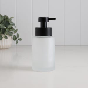 May include: A frosted glass soap dispenser with a black pump. The dispenser has a cylindrical shape and is on a white surface. The background is a white tiled wall.