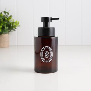 May include: A brown glass soap dispenser with a black pump. The dispenser has a white label with the words "HAND SOAP" in a circular design. The dispenser is on a white surface.